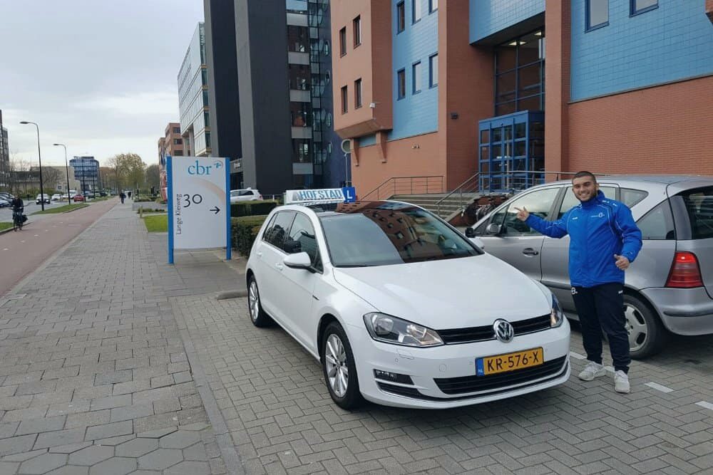Geslaagde leerling met duimen omhoog bij de witte lesauto van Hofstad Rijopleiding, bij het CBR in Rijswijk